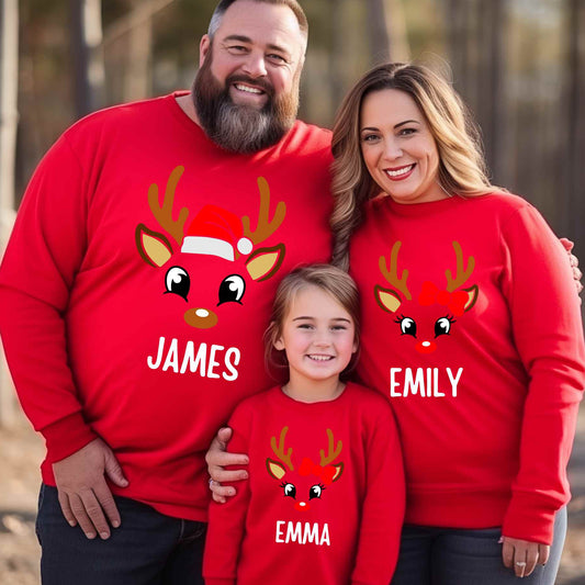 Family of three wearing red Christmas-themed sweaters with reindeer designs and names.