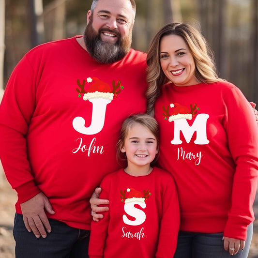Family of three wearing red Christmas-themed sweaters with personalized names outdoors.